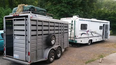 A stock trailer and an RV parked on gravel with trees behind.